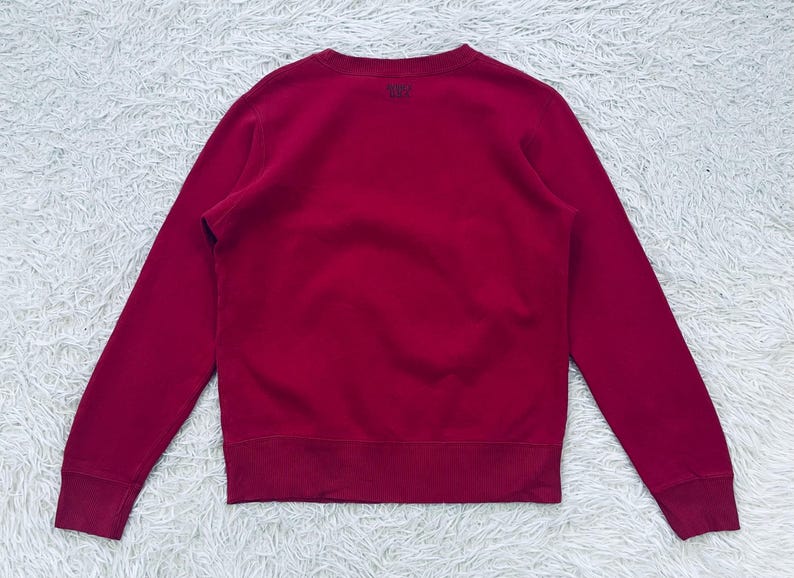 May include: A solid crimson sweatshirt with a crew neck and long sleeves. The garment has ribbed cuffs and a ribbed hem. A small logo is visible on the upper back. The sweatshirt is made of a soft, comfortable material.