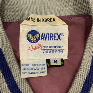 K&ouml;nnte beinhalten: Nahaufnahme eines Jackenetiketts mit dem Text "MADE IN KOREA" und der Marke "AVIREX&reg;". Das Etikett enth&auml;lt auch "Varsity CLUB MEMBERSHIP" und die Gr&ouml;&szlig;e "M". Die Jacke ist beige mit blauen Streifen.