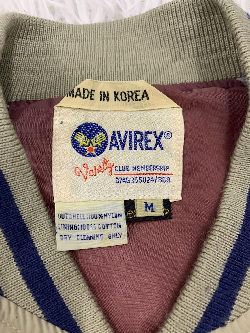 K&ouml;nnte beinhalten: Nahaufnahme eines Jackenetiketts mit dem Text "MADE IN KOREA" und der Marke "AVIREX&reg;". Das Etikett enth&auml;lt auch "Varsity CLUB MEMBERSHIP" und die Gr&ouml;&szlig;e "M". Die Jacke ist beige mit blauen Streifen.