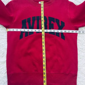 May include: A red sweatshirt with the word "AVIREX" in dark green block letters across the chest. The sweatshirt has a crew neck and ribbed cuffs and hem. A measuring tape is laid across the sweatshirt.