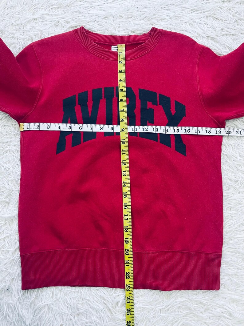 May include: A red sweatshirt with the word "AVIREX" in dark green block letters across the chest. The sweatshirt has a crew neck and ribbed cuffs and hem. A measuring tape is laid across the sweatshirt.