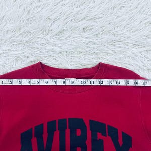 May include: A red sweatshirt with the word "AVIREX" in navy blue block letters across the chest. A measuring tape is placed across the top of the sweatshirt, showing a measurement of approximately 20 inches.