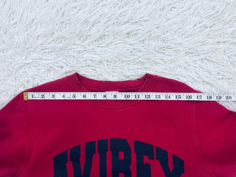 May include: A red sweatshirt with the word "AVIREX" in navy blue block letters across the chest. A measuring tape is placed across the top of the sweatshirt, showing a measurement of approximately 20 inches.