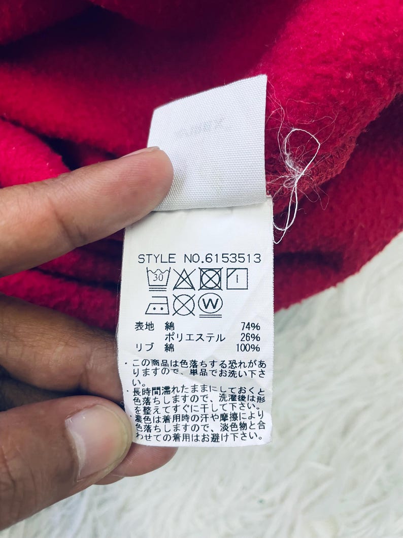 May include: Close-up of a red fabric with a white tag attached. The tag displays washing instructions and fabric composition details, including 74% cotton and 26% polyester. The style number is 6153513.
