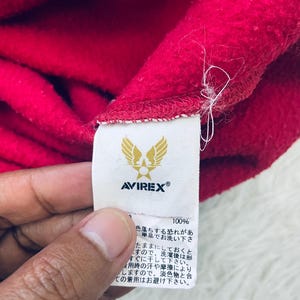 May include: Close-up of a red fleece fabric with a white tag. The tag features the brand name "AVIREX" and a logo of a bird with spread wings. The tag also includes Japanese text and the number "100%."