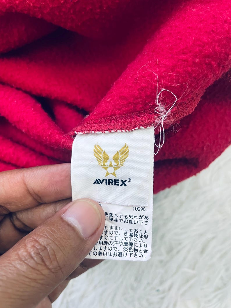 May include: Close-up of a red fleece fabric with a white tag. The tag features the brand name "AVIREX" and a logo of a bird with spread wings. The tag also includes Japanese text and the number "100%."