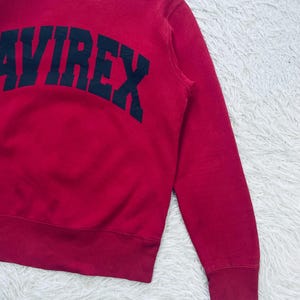 May include: A red sweatshirt with the word "AVIREX" printed in navy blue across the chest. The sweatshirt has a crew neck and ribbed cuffs and hem. The fabric appears to be a soft, comfortable material.