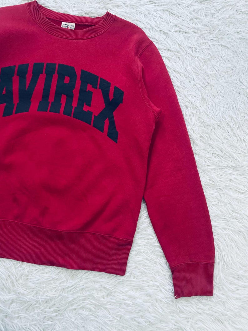 May include: A red sweatshirt with the word "AVIREX" printed in navy blue across the chest. The sweatshirt has a crew neck and ribbed cuffs and hem. The fabric appears to be a soft, comfortable material.