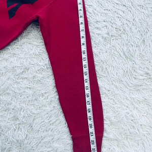 May include: A red sweatshirt with the word "EX" in black block letters. The sleeve is measured with a tape measure, showing a length of approximately 24 inches.
