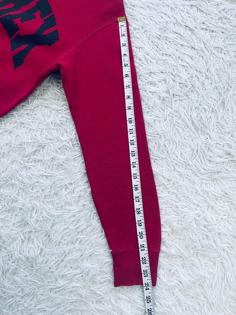 May include: A red sweatshirt with the word "EX" in black block letters. The sleeve is measured with a tape measure, showing a length of approximately 24 inches.