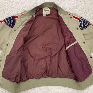 K&ouml;nnte beinhalten: Beige Bomberjacke mit roten und blauen Aufn&auml;hern. Die Jacke hat ein bordeauxfarbenes Futter und einen gerippten Kragen und B&uuml;ndchen. Aufn&auml;her enthalten Text und Grafiken. Die Jacke hat eine Knopfleiste und zwei Taschen.