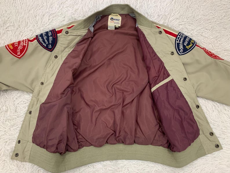 K&ouml;nnte beinhalten: Beige Bomberjacke mit roten und blauen Aufn&auml;hern. Die Jacke hat ein bordeauxfarbenes Futter und einen gerippten Kragen und B&uuml;ndchen. Aufn&auml;her enthalten Text und Grafiken. Die Jacke hat eine Knopfleiste und zwei Taschen.