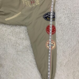 K&ouml;nnte beinhalten: Beige Bomberjacke mit gestickten Aufn&auml;hern und Text. Die Jacke hat eine gestreifte Manschette und ein Ma&szlig;band, das eine L&auml;nge von etwa 84 cm anzeigt.