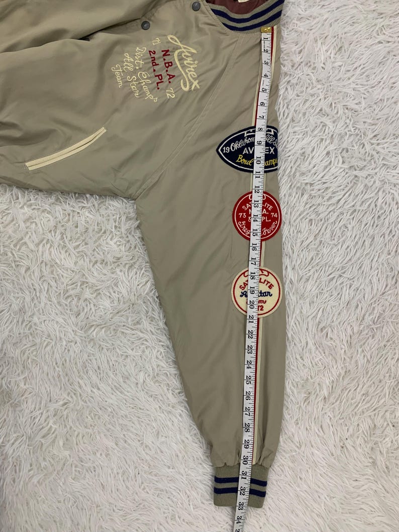 K&ouml;nnte beinhalten: Beige Bomberjacke mit gestickten Aufn&auml;hern und Text. Die Jacke hat eine gestreifte Manschette und ein Ma&szlig;band, das eine L&auml;nge von etwa 84 cm anzeigt.
