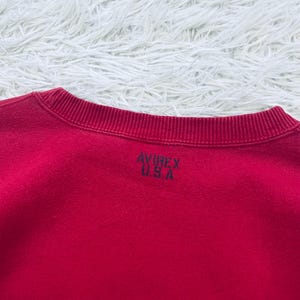 May include: A red crew neck sweatshirt with the text "AVIREX U.S.A." printed in black on the back. The sweatshirt is made of a soft material and has a ribbed neckline.