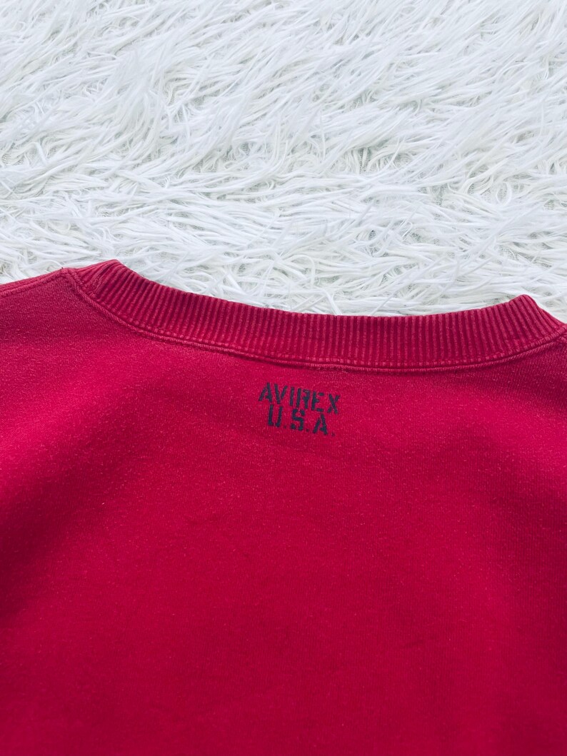 May include: A red crew neck sweatshirt with the text "AVIREX U.S.A." printed in black on the back. The sweatshirt is made of a soft material and has a ribbed neckline.