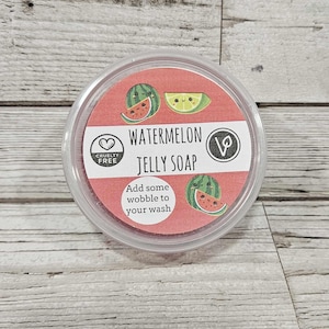 May include: A clear plastic container with a red lid containing watermelon jelly soap. The label says "Watermelon Jelly Soap" and features a watermelon and lime design. The label also says "Cruelty Free" and "Vegan".