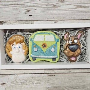 May include: A white box containing three bath bombs. The bath bombs are shaped like Scooby Doo, the Mystery Machine van, and Shaggy. The bath bombs are on a bed of shredded paper.