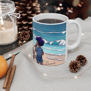 Könnte beinhalten: Eine weiße Keramiktasse mit einer Cartoon-Illustration einer Person, die an einem Strand mit blauen Wellen und einem sandigen Ufer steht. Die Tasse hat einen schwarzen Henkel und ist mit einer dunklen Flüssigkeit gefüllt.