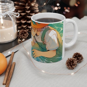 Könnte beinhalten: Weiße Keramiktasse mit einer bunten Illustration einer Person, die einen Strohhut und eine Latzhose trägt und im Wasser steht. Die Tasse hat einen Henkel auf der rechten Seite.