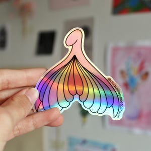 Può includere: Adesivo olografico con un design stilizzato e astratto nei toni del rosa, giallo e blu. L'adesivo ha una forma unica con una base curva e una parte superiore arrotondata. Il testo "@coquillagesetclitoris" è stampato sull'adesivo.