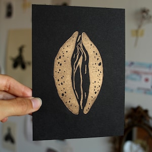 Könnte beinhalten: Eine schwarze Karte mit einer goldfarbenen Illustration einer Vulva. Das Kunstwerk hat ein strukturiertes Aussehen mit kleinen, dunklen Punkten. Das Bild wird von einer Hand gehalten, wobei andere Kunstwerke im Hintergrund sichtbar sind.
