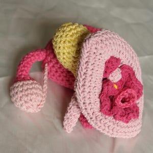 May include: A handcrafted crochet toy featuring a soft pink hat with a dark pink flower accent, a bright yellow rounded top section, and a dangling pink arm with a light pink spherical hand, all constructed from textured yarn stitches.
