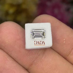 Könnte beinhalten: Ein Diamant im Smaragdschliff auf einer weißen Präsentationskarte. Der Diamant ist rechteckig mit einer Stufenschliff-Anordnung, die Licht reflektiert. Auf der Karte steht der Text "DADA DIAMOND JEWELLERY" in Rot. Der Hintergrund ist verschwommen mit floralen Farben.
