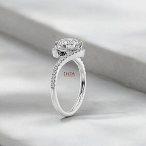 May include: A silver engagement ring with a large, round, clear gemstone at the center. The band twists and is encrusted with smaller, clear gemstones. The word "DADA" is visible on the ring.