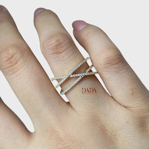 Op de afbeelding: Een goudkleurige ring met een kruisontwerp, bezet met kleine, heldere stenen langs de banden. De ring zit om een vinger, met de merknaam "DADA" zichtbaar. Het ontwerp van de ring is modern en elegant.