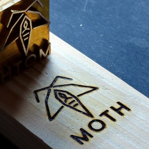 Puede incluir: Un bloque de madera con la palabra "MOTH" grabada. Un sello de latón con un diseño de polilla se coloca sobre la madera. El diseño es una polilla estilizada con una forma ovalada central. El fondo es oscuro.