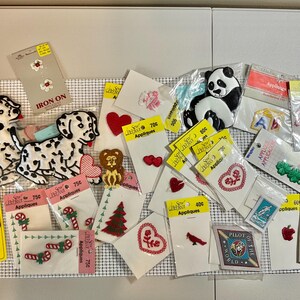 May include: A collection of various iron-on fabric appliques in individual retail packaging. Designs include dalmatian dogs, a panda, a teddy bear, hearts, candy canes, a Christmas tree, a lion, an apple, and a Pilot of the World emblem.