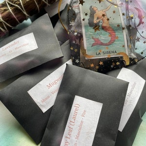 May include: A collection of small black pouches with white labels, each containing a different type of herb. The labels list the herb name, its astrological association, and whether it is masculine or feminine. The pouches are arranged on a surface with a piece of dried sage and a small bag containing a tarot card with the image of a mermaid.