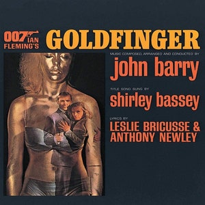 May include: A vintage record album cover for the James Bond film "Goldfinger". The cover features a woman in a gold bikini with two men behind her. The title "Goldfinger" is in large yellow letters. The album is by John Barry, with the title song sung by Shirley Bassey. The lyrics are by Leslie Bricusse and Anthony Newley.