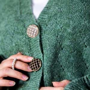 May include: A green cardigan with two decorative buttons. The buttons are round and have a grid pattern. One button is white with gold trim and the other is black with gold trim.
