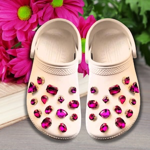 May include: A pair of white clogs decorated with pink rhinestones. The clogs are sitting on a wooden surface with pink flowers in the background.