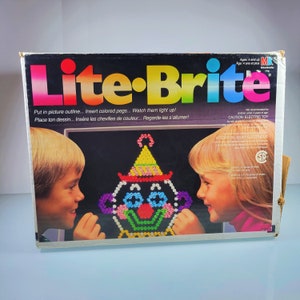 May include: A Lite-Brite toy box with a colorful clown image on the front. The box has the text "Lite-Brite" in large, colorful letters. The text "Put in picture outline... Insert colored pegs... Watch them light up!" is printed below the Lite-Brite logo. The text "Place ton dessin... Insère les chevilles de couleur... Regarde-les s'allumer!" is printed below the first line of text. The box is white with black trim. The box is for ages 4 and up.