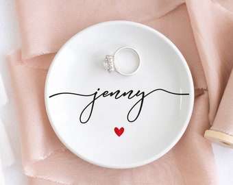 May include: White ceramic ring dish with the name "jenny" written in black cursive script and a small red heart. A silver ring with a clear stone is sitting on the dish.