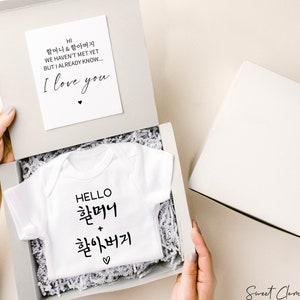 May include: A white onesie with the text "HELLO 할머니 + 할아버지" in black. The onesie is in a white gift box with white tissue paper. A card with the text "HI 할머니 & 할아버지 WE HAVEN'T MET YET BUT I ALREADY KNOW... I love you" is visible in the background.