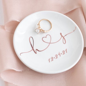 May include: White ceramic ring dish with a rose gold inscription that reads "hers" with a heart and the date "12.24.21". A gold ring with a large clear stone is sitting on the dish.