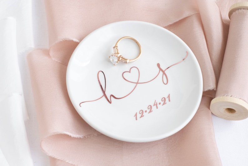 May include: White ceramic ring dish with a rose gold inscription that reads "hers" with a heart and the date "12.24.21". A gold ring with a large clear stone is sitting on the dish.