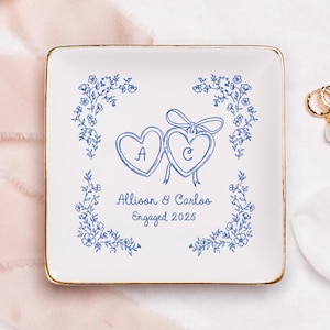 Pode incluir: Prato quadrado de cerâmica branca com borda dourada, apresentando um design floral azul e dois corações conectados com as letras "A" e "C". O texto "Allison & Carlos Engaged 2025" também é exibido.