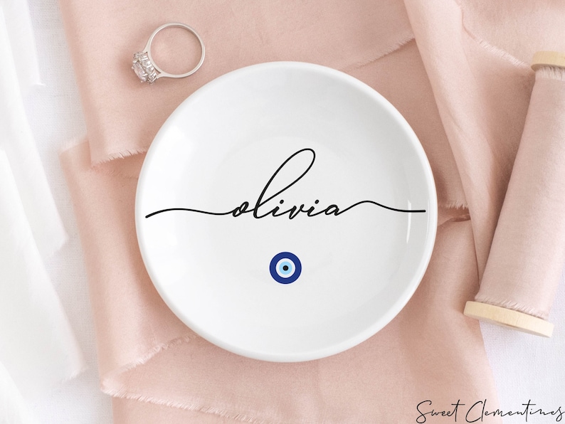 May include: White ceramic ring dish with the name "Olivia" written in black cursive script. A blue and white evil eye charm is centered below the name.