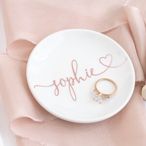 May include: A white ceramic ring dish with the name "Sophie" written in pink script and a pink heart. A gold ring with a large diamond is sitting on the dish.