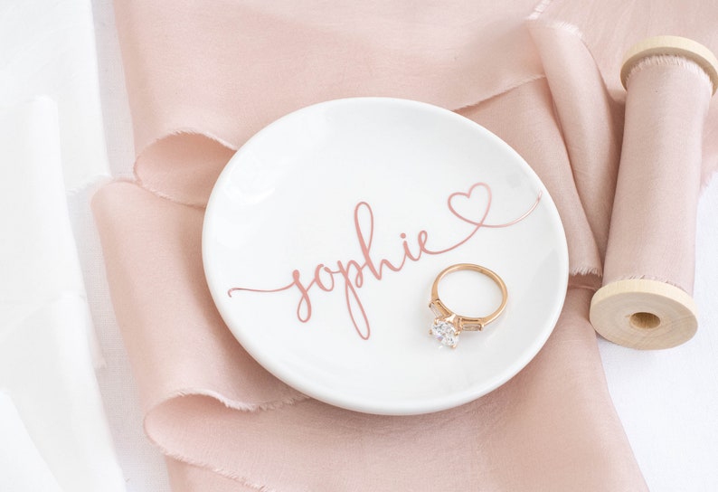 May include: A white ceramic ring dish with the name "Sophie" written in pink script and a pink heart. A gold ring with a large diamond is sitting on the dish.