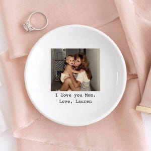 以下が含まれることがあります： 母と子の写真がプリントされた白いセラミック製のリングトレイ。「I love you Mom. Love, Lauren」の文字が入っています。大きな透明な石が付いたシルバーの指輪がトレイの近くに置かれています。