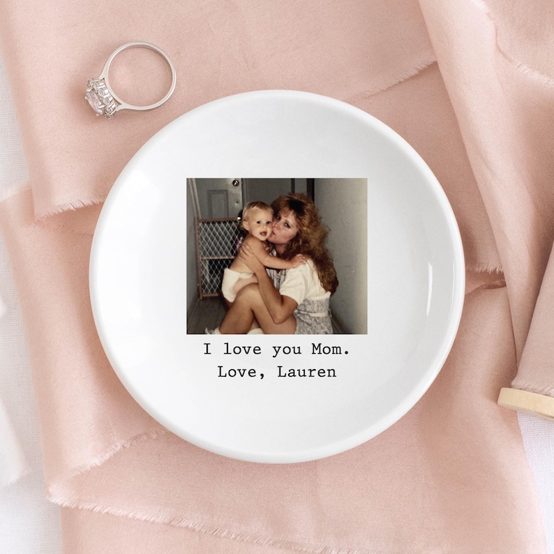 以下が含まれることがあります： 母と子の写真がプリントされた白いセラミック製のリングトレイ。「I love you Mom. Love, Lauren」の文字が入っています。大きな透明な石が付いたシルバーの指輪がトレイの近くに置かれています。