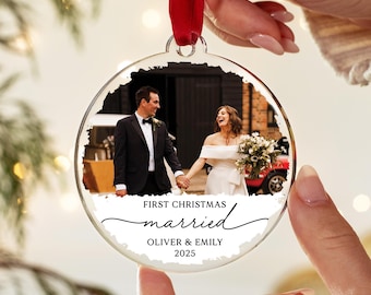 May include: Clear, round Christmas ornament with a wedding photo inside. The photo shows a couple holding hands. Text on the ornament reads "First Christmas Married" with the names "Oliver & Emily" and the year "2025". A red ribbon is attached.