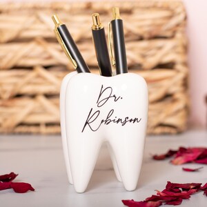 May include: White ceramic tooth-shaped desk organizer with the text "Dr. Robinson" in black script. Three black pens are in the organizer.