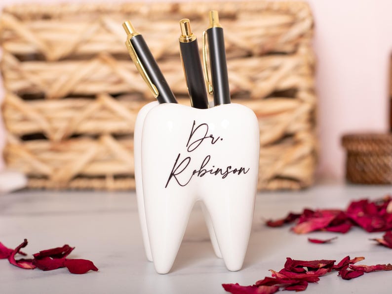 May include: White ceramic tooth-shaped desk organizer with the text "Dr. Robinson" in black script. Three black pens are in the organizer.